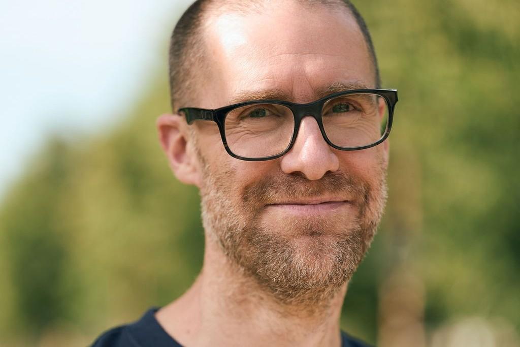 Portret van een man met bril en kort haar, buiten gefotografeerd met bomen op de achtergrond.