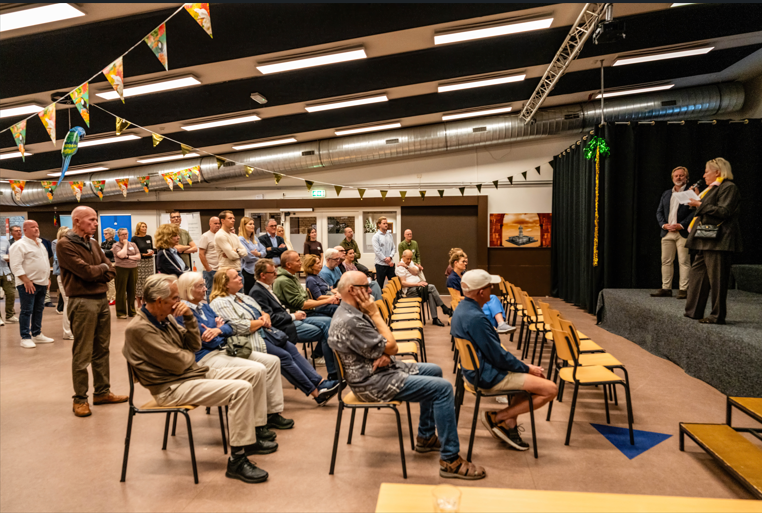 Zaal met mensen die luisteren naar wethouder op podium
