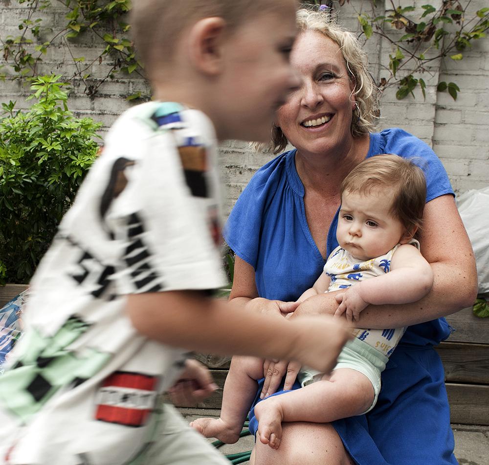 Foto van vrouw in blauwe jurk met baby op schoot en jongetje dat voorbij rent