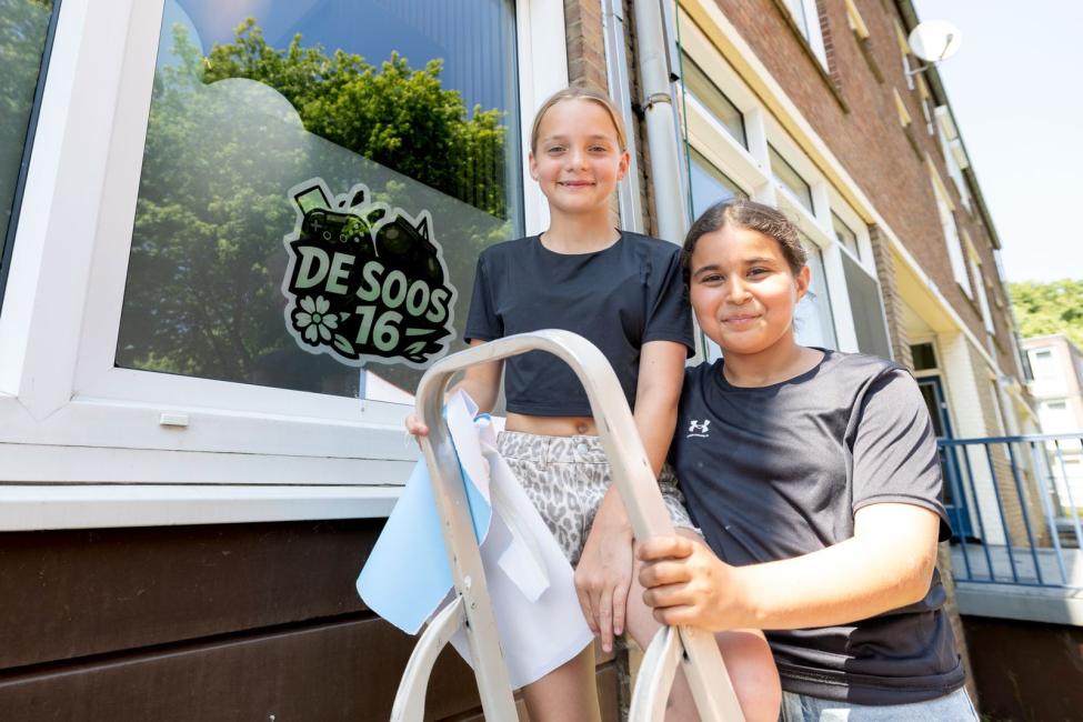Twee meisjes staan bij het raam van jeugdsoos De Soos 16 en houden een ladder vast.
